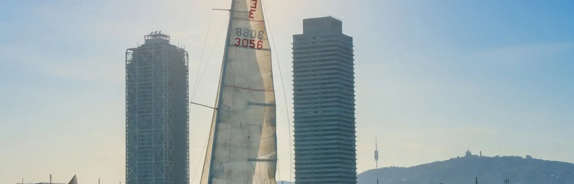 Veler navegant davant de l'skyline de Barcelona amb el sol baix i la mar en calma