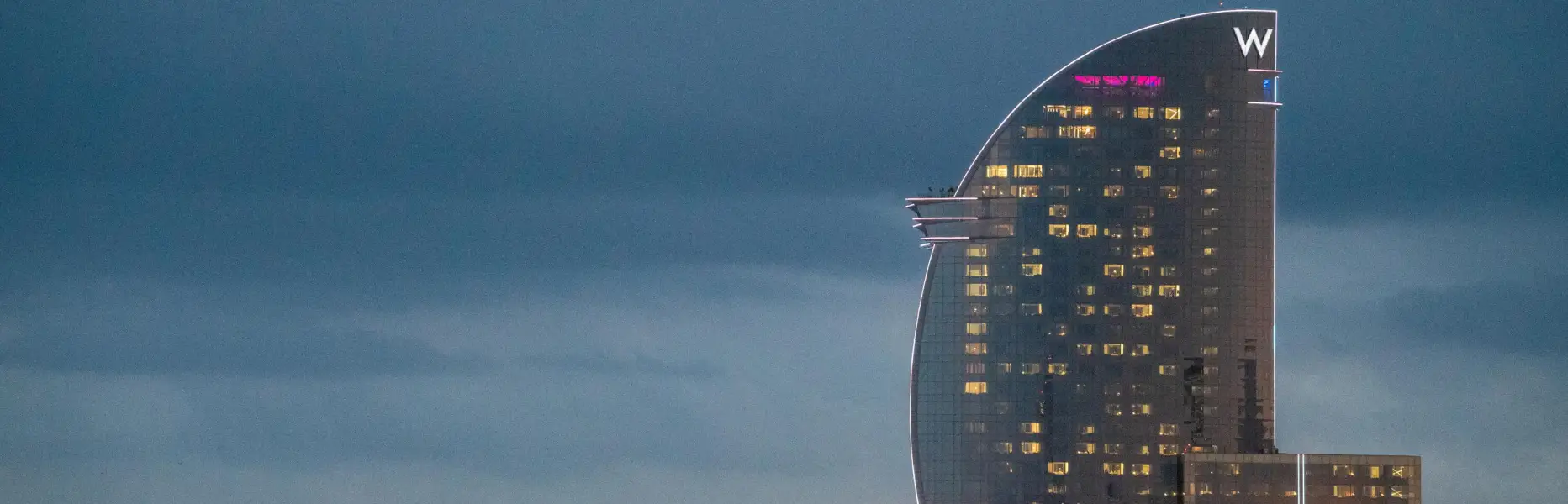 Blick auf das W Hotel in Barcelona bei Sonnenuntergang am Meer mit eingeschalteten Lichtern und bewölktem Himmel