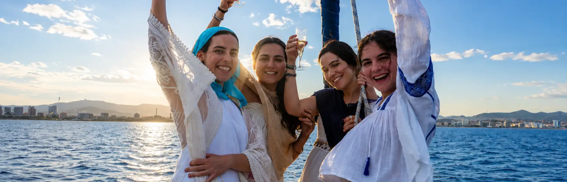 Groupe d’amies trinquant au cava à la proue d’un voilier privé face à la côte de Barcelone au coucher du soleil