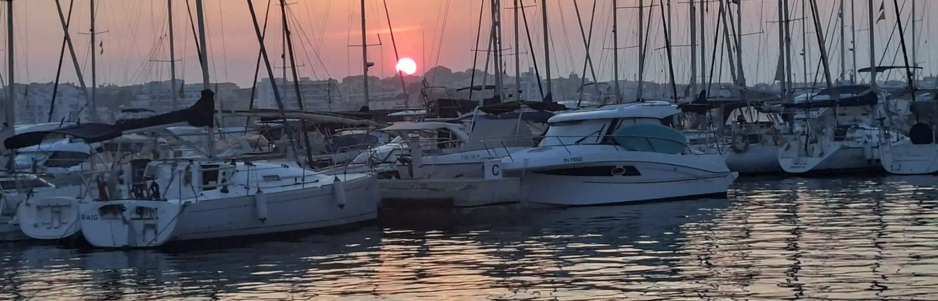 Barca a vela e yacht ormeggiati in un porto di Barcellona al tramonto con il sole sull’orizzonte e il mare calmo