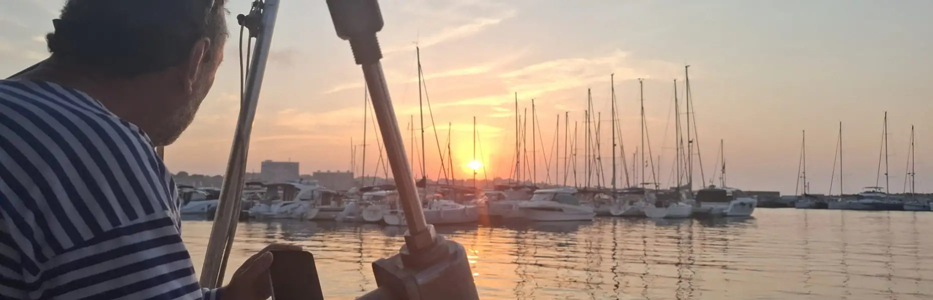 Skipper su barca a vela privata che osserva il tramonto sul porto turistico di Barcellona con numerose barche a vela ormeggiate