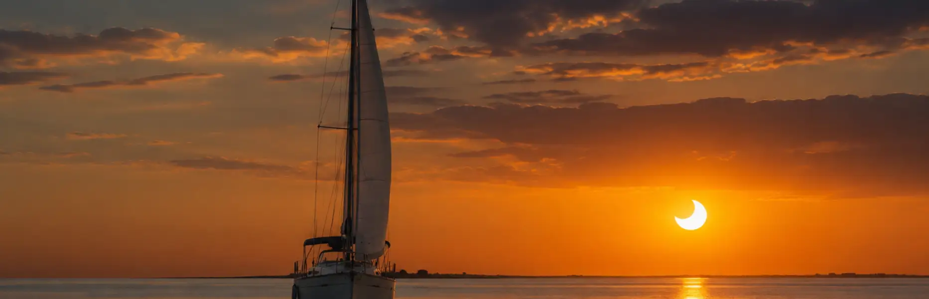 Velero navegando al atardecer sobre un mar en calma mientras el Sol muestra un eclipse parcial tras las nubes