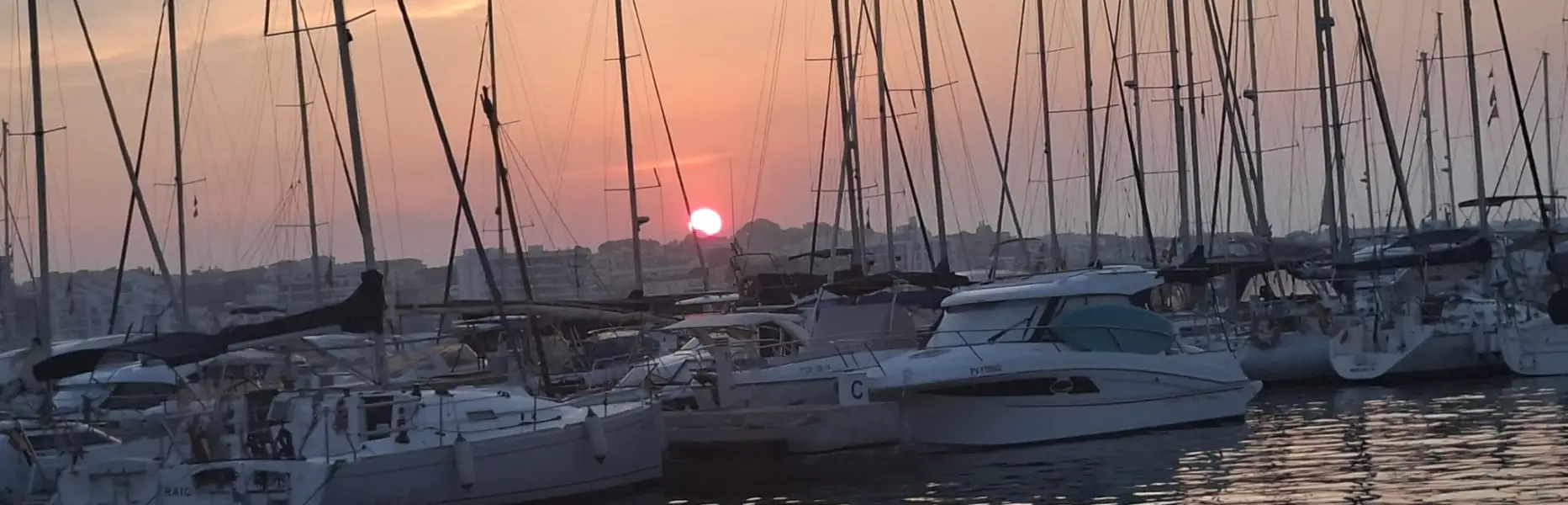 Pink sunset over a harbour with moored sailboats and the sun reflecting on the sea in Barcelona